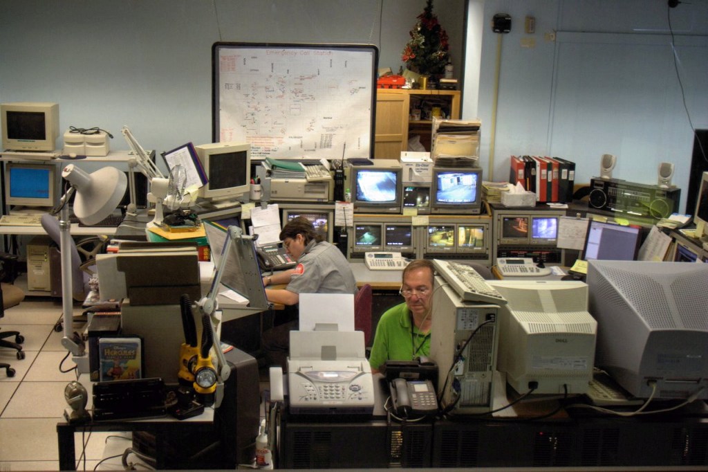 Night-time view through a window into a security room where staff watch banks of surveillance monitors.